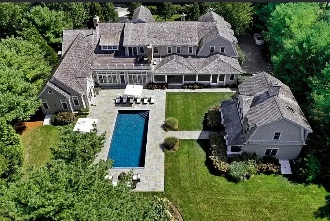 Aerial view of a large shingle-style house with a rectangular pool, stone terrace and clipped lawn, surrounded by mature trees.