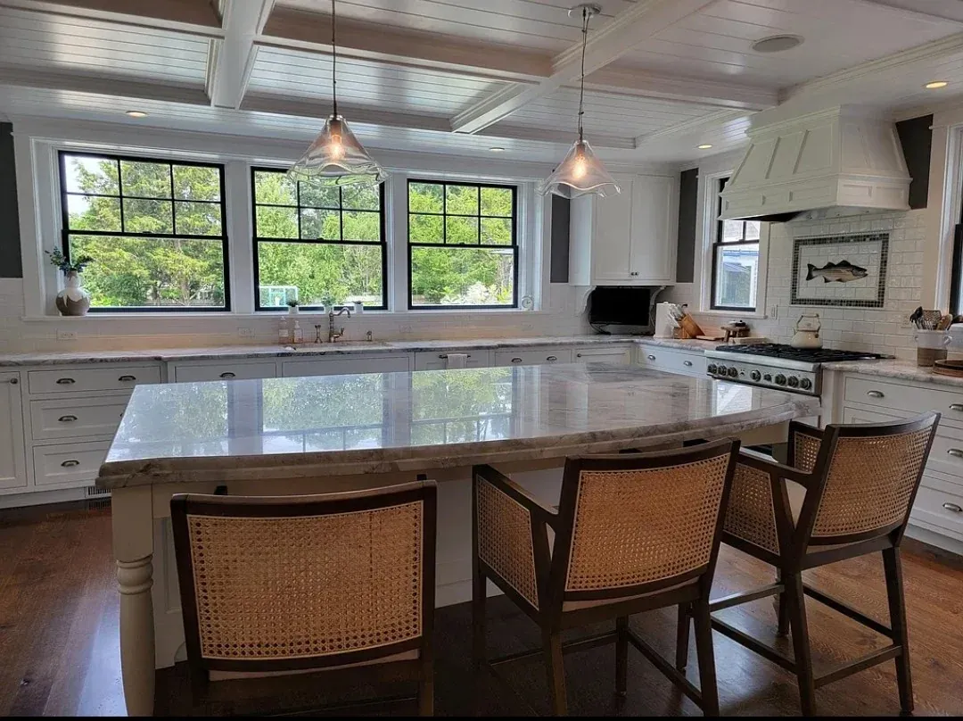 A white kitchen with a coffered plank ceiling, a long marble island with cane bar stools and a row of black-framed windows.
