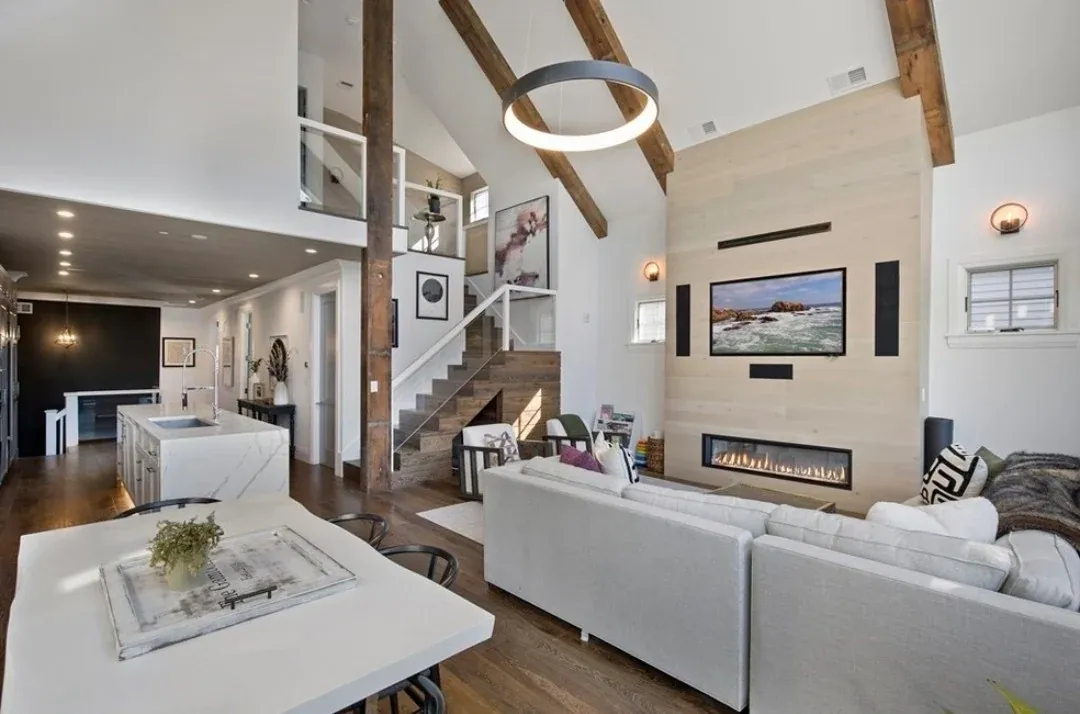 An open-plan living room with exposed timber beams, a vaulted white ceiling, a linear gas fireplace and a staircase behind.