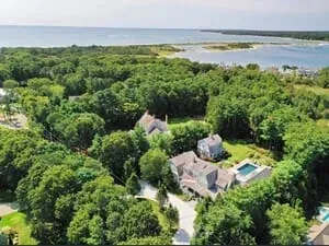 Aerial view over a wooded Cape neighbourhood, a pool and a shingled house in the foreground and an inlet beyond.