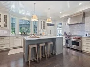 A white kitchen with a large island, hardwood floor, pendant lights and a stainless range under a hood.