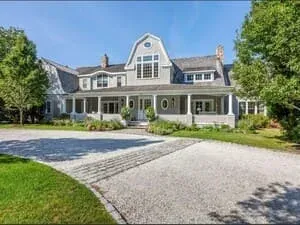 A large shingle-style house with a covered porch, dormers and a gravel turning circle.