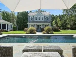 A rectangular pool with a market umbrella and lounge chairs, the house and lawn behind.