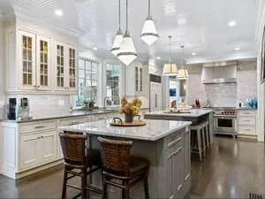 A white kitchen with two islands, glass-front upper cabinets and a run of pendant lights.