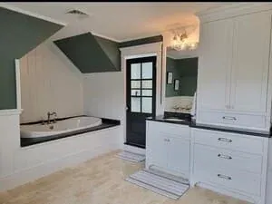 A bathroom with a drop-in tub under a sloped ceiling, white cabinetry and a dark glazed door.