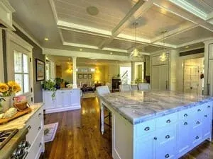 A kitchen with a coffered ceiling, a blue painted island and a marble counter running to a bright dining end.