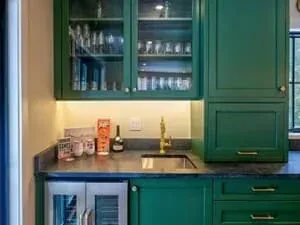 A bar with deep green cabinetry, glass-front uppers, a dark stone counter and glassware on the shelves.