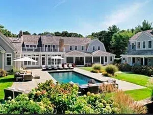 Aerial view of a shingled estate with a pool, cabana and loungers set into a hedged lawn.