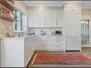 A white kitchen with a patterned red runner, a window over the sink and glass-front cabinets.