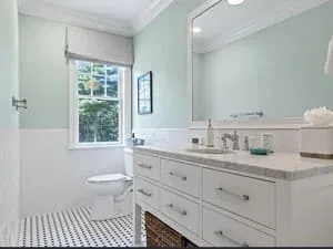 A bathroom with a white double vanity, a marble top and a window above the tub.