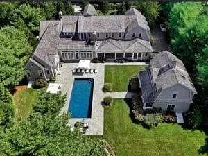 Aerial view of a gambrel-roofed shingled house with a pool set into a hedged garden.