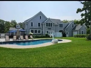 A shingled house with a pool, lounge chairs and a wide lawn running to the treeline.
