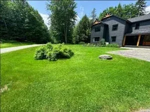 A wide lawn with a boulder in the foreground and a dark shingled house beyond.