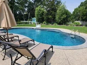 A pool with lounge chairs and a folded umbrella on a paved surround beside a hedge.