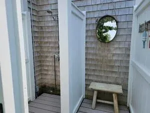 A cedar-shingled outdoor shower enclosure with a round mirror and a teak bench.