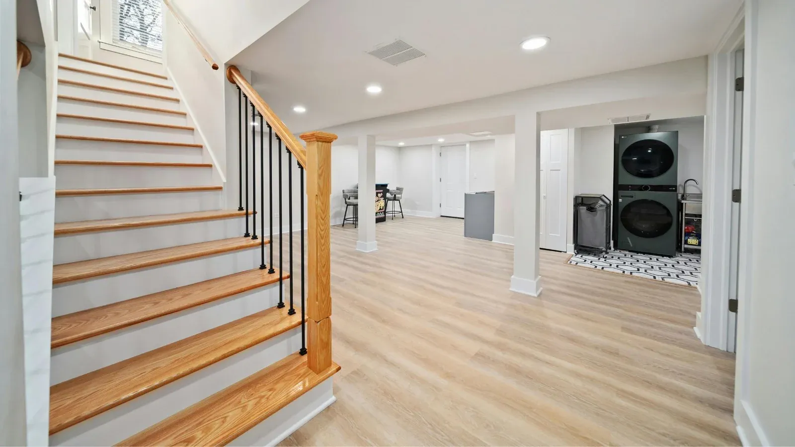 A white oak staircase with black metal balusters beside a bright laundry area with two stacked machines and a tiled floor.